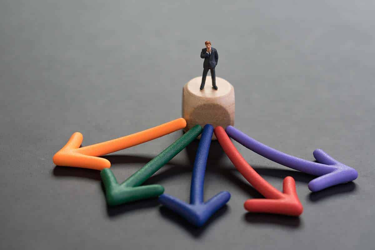 Miniature businessman man standing and thinking on wooden cube with multi direction color arrow pointing out on blackboard.